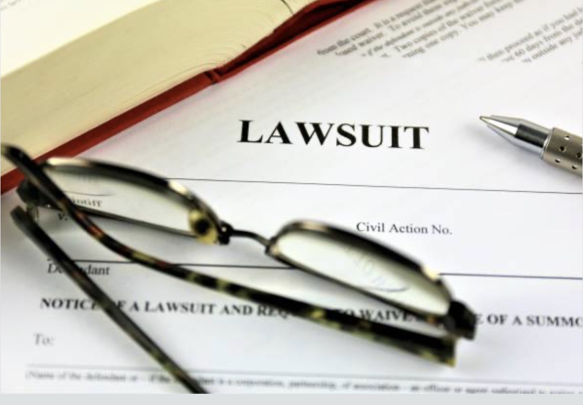 Close-up of a legal document titled "LAWSUIT" with reading glasses resting on top. A pen and an open book are visible, suggesting a legal context.