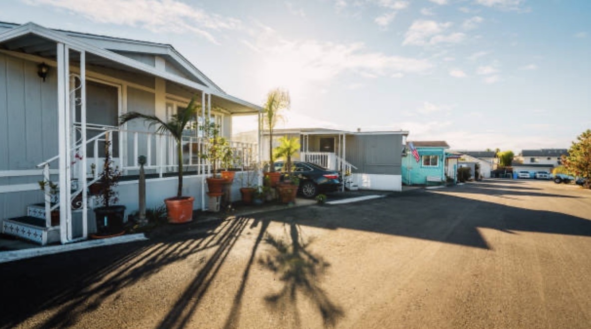 A sunny day in a mobile home park shows tidy homes with potted plants, a parked car, and palm tree shadows on the pavement, creating a serene atmosphere.