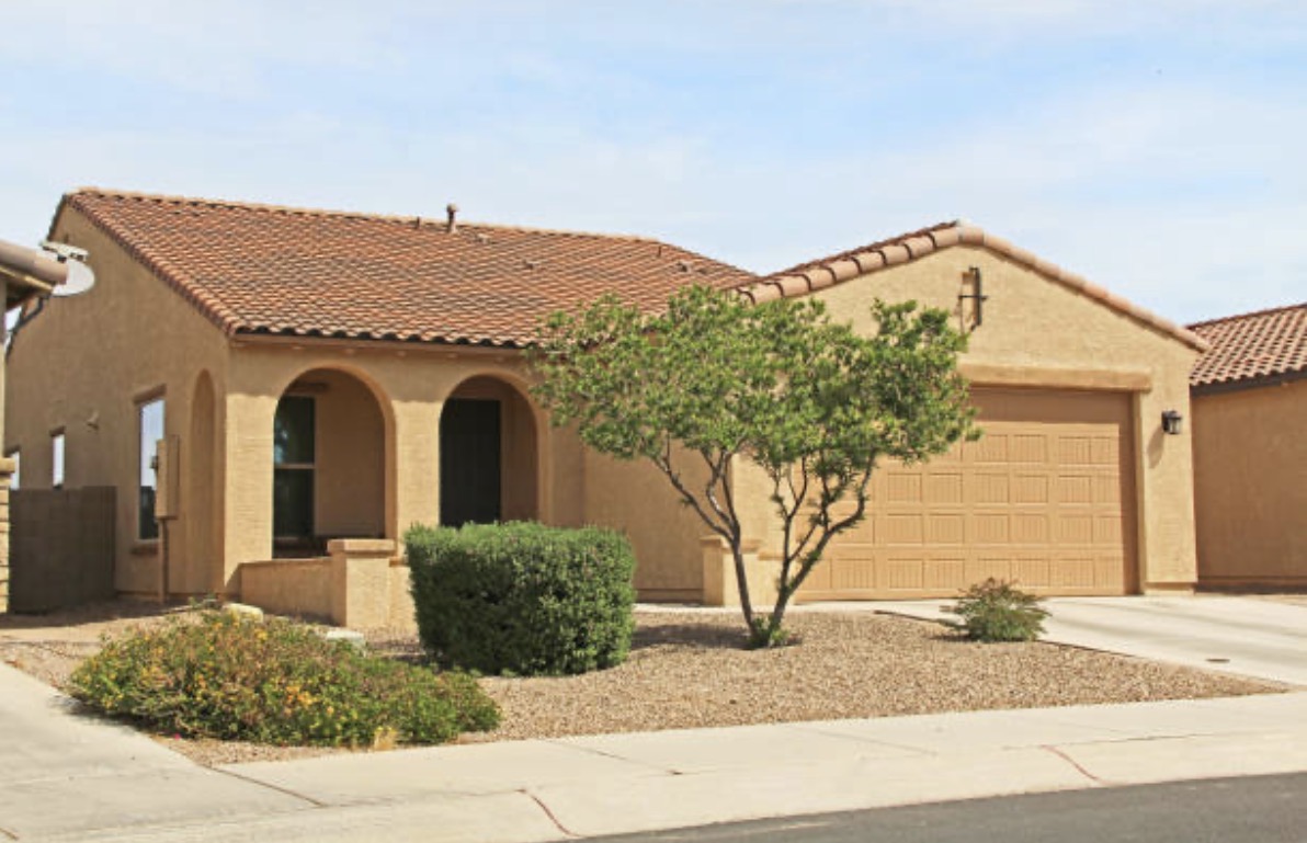 Single-story house with a terracotta-tiled roof and stucco exterior. Features arched entryway, two-car garage, and desert landscaping. Bright, sunny day.