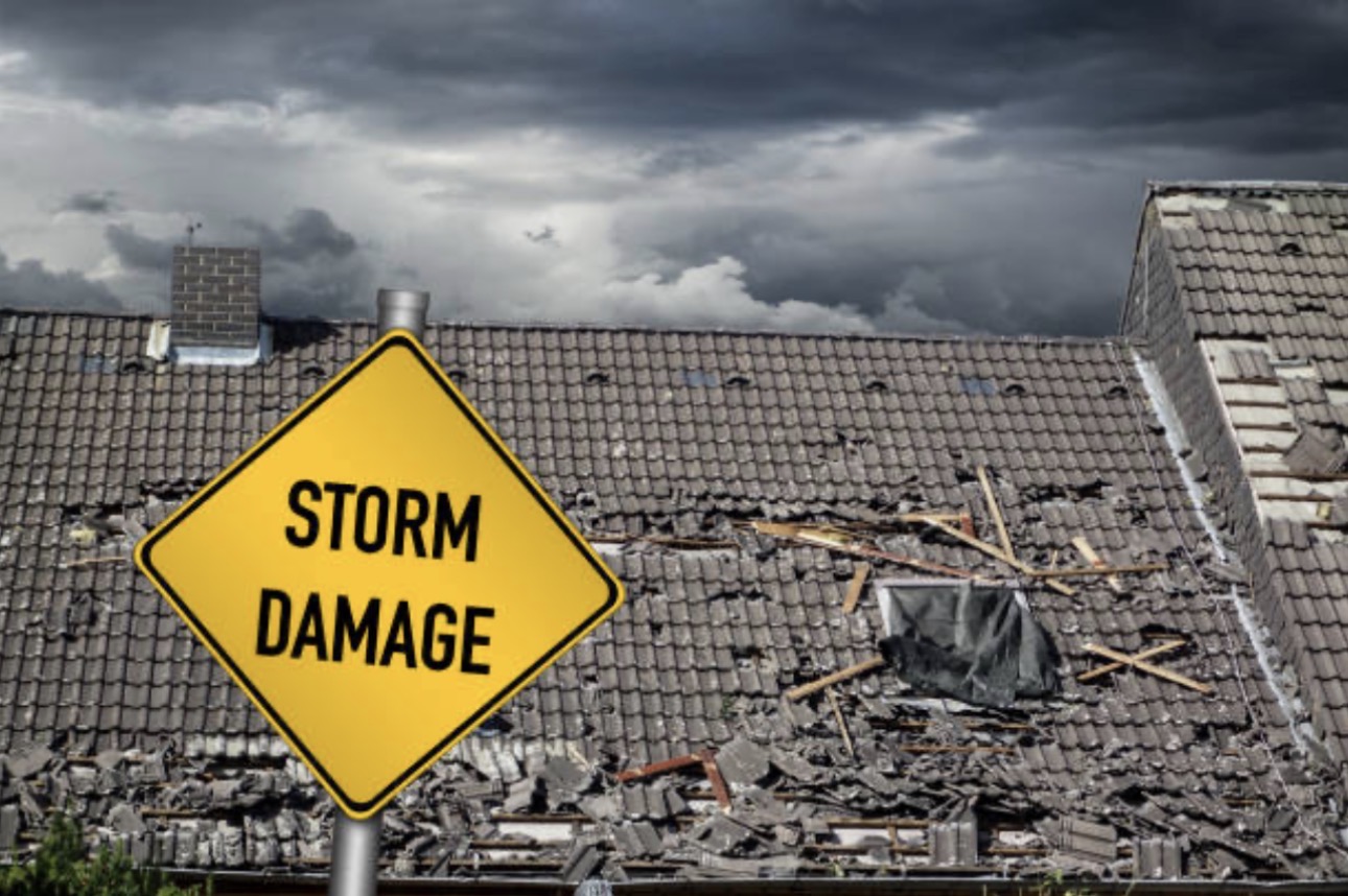 A damaged roof with missing tiles is seen under dark, stormy clouds. A yellow sign in the foreground reads "Storm Damage," highlighting the destruction.