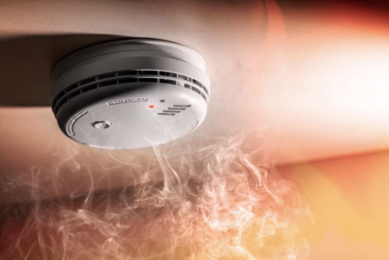 Smoke alarm on ceiling with red indicator light glowing, surrounded by smoke and a warm glow, suggesting a fire alert scenario.