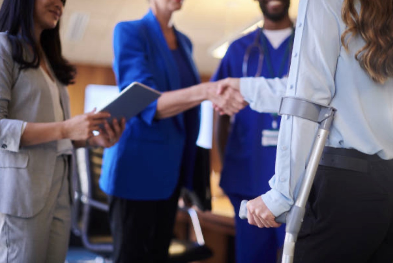 A person on crutches shakes hands with a doctor in a hospital meeting room, surrounded by others in formal attire, creating a supportive atmosphere.
