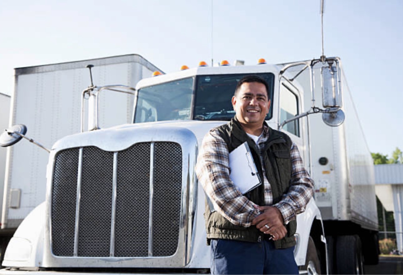 Man smiling confidently in front of a large white semi-truck, holding a clipboard. He wears a plaid shirt and vest, conveying professionalism.