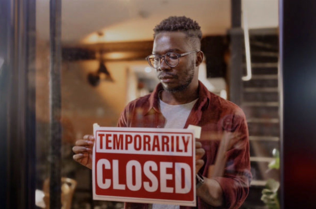 A man in a brown jacket holds a "Temporarily Closed" sign inside a dimly lit shop. The atmosphere is somber and reflective.